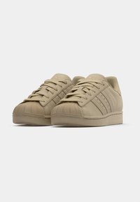 Beige sneakers with a textured toe cap, flat lace-up design, three perforated stripes on the side, and a padded collar.