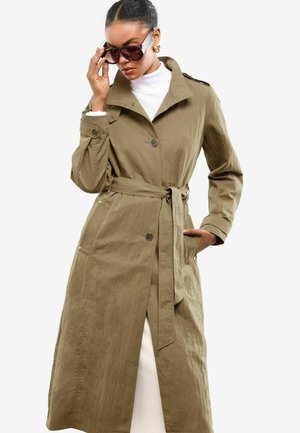 Jonge vrouw in een lichtbruin trenchcoat en donkere zonnebril, die haar bril aanpast terwijl ze zelfverzekerd poseert tegen een witte achtergrond.