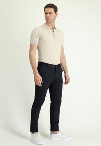 Beżowa koszulka polo z krótkim rękawem i detalem przy kołnierzyku na zamek, zestawiona z czarnymi spodniami typu slim fit i białymi sneakersami, na neutralnym tle.