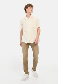 Lichtbeige overhemd met korte mouwen, een zak, verticale textuur en knoopsluiting, gecombineerd met lichtbruine slim-fit broeken en witte sneakers.