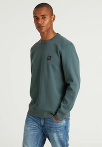 Sudadera verde de tela suave con escote redondo, que presenta un pequeño parche de logotipo negro en el pecho y puños acanalados.