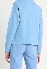 Vaaleansininen ylisuurikokoinen neulepaita, jossa on korkea kaulus ja pitkät hihat. Neulepaidan pinta on sileä, ja siinä on hieman laskeutuvat hihat. Tummansiniset housut.