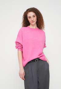 Γυναίκα με σγουρά κόκκινα μαλλιά, φοράει ένα φωτεινό ροζ πουλόβερ και γκρι πλισέ παντελόνι, στέκεται με το ένα χέρι στην τσέπη μπροστά σε λευκό φόντο.