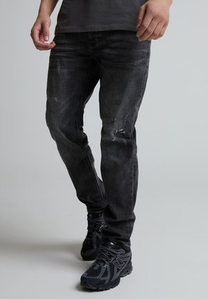 Man met verweerde zwarte jeans met kleine scheuren en zwarte sportschoenen, staand tegen een effen lichte achtergrond.