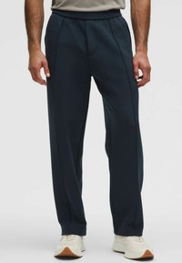 Mørkeblå sportbukser i blødt, strækbart stof med et strømlinet design, plisseret front og elastisk talje, parret med hvide sneakers.