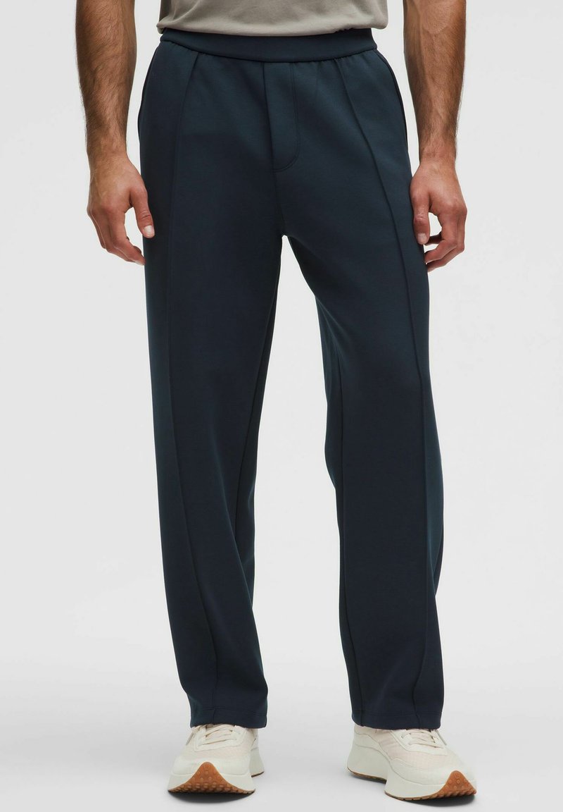 Pantalones deportivos azul marino de tejido suave y elástico, con un diseño aerodinámico, fruncido en la parte delantera y cinturilla elástica, combinados con zapatillas blancas.