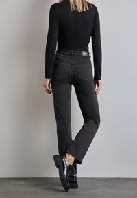 Jeans de denim negro con un diseño de pierna recta, que presentan un ligero desgaste, bolsillos traseros y una etiqueta en la cinturilla. Estilizados con zapatos negros.