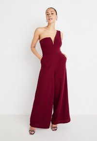 Jumpsuit bordeaux con design a spalla unica, scollatura a V profonda, gamba larga e tasche laterali, realizzato in tessuto morbido e liscio.
