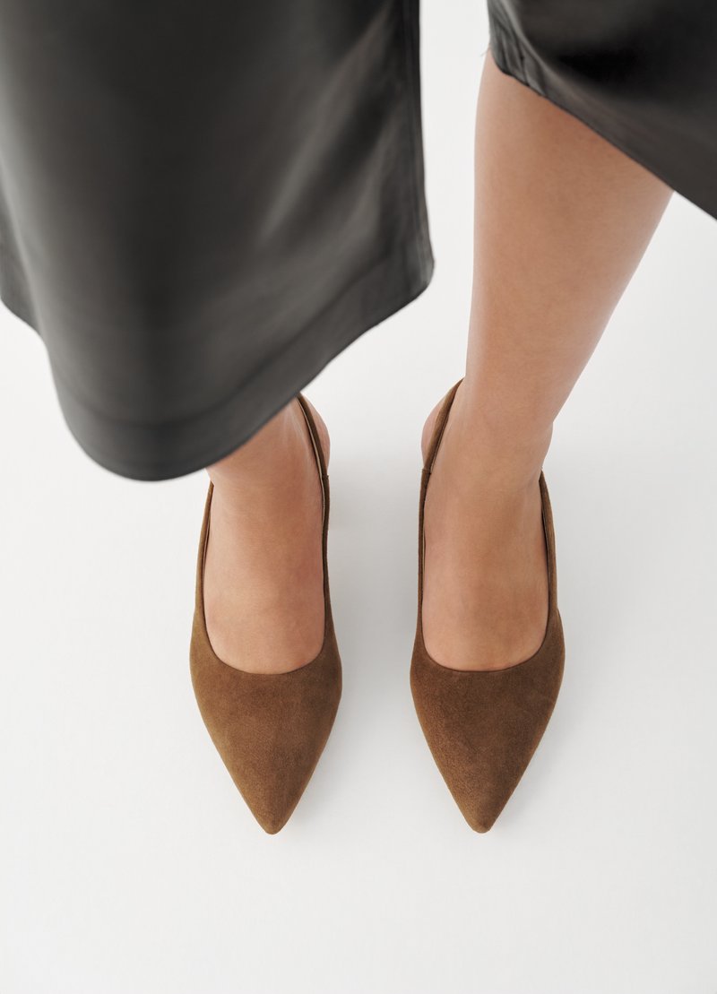 Füße, die braune Wildleder-Pumps mit spitzer Kappe und Slingback tragen, teilweise bedeckt von einem schwarzen Rock auf weißem Hintergrund.