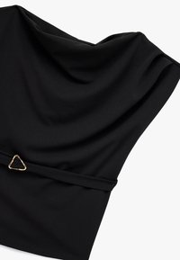 Czarna tkanina bluzki ma drapowany fason z trójkątnym złotym akcentem w talii, gładką fakturę i luźny krój.