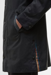 Casaco impermeável preto com botões de pressão, bolsos laterais e um detalhe em riscas de arco-íris ao longo da costura. Textura suave com um corte moderno.