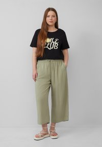 T-shirt noir avec le texte "Smile Club", pantalon large olive, et sandales à lanières de couleur claire. Design simple, coupe décontractée et texture lisse.