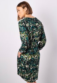 Grün gemustertes Kleid mit langen Ärmeln und eingezogenem Bund, das Taschen und ein lockeres, fließendes Design aufweist. Das Muster umfasst verschiedene Grüntöne, Schwarz und Weiß.