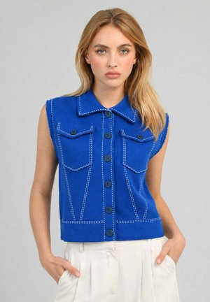 Femme portant un haut bleu sans manches boutonné avec des coutures blanches et un pantalon blanc taille haute, debout les mains dans les poches devant un fond gris.