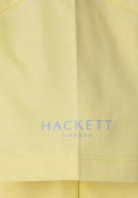 Żółta bawełniana tkanina o gładkiej fakturze, z napisem "HACKETT LONDON" w jasnoniebieskim kolorze, umieszczonym w dolnej części.