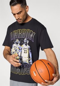 Μαύρο βαμβακερό μπλουζάκι με πολύχρωμο γραφικό ενός παίκτη μπάσκετ, με το κείμενο "LEBRON JAMES", συνοδευόμενο από πορτοκαλί μπάλα μπάσκετ.