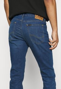 Man med mörkblå Lee-jeans och svart skjorta, sedd från midjan och neråt med en hand vilande nära höften.