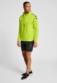 Heldere limoengroene hoodie met trekkoorden en zwarte chevron accenten, gecombineerd met zwarte shorts. Model draagt zwarte sportschoenen.