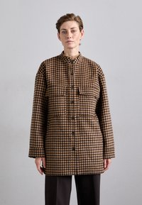 Een bruin en zwart houndstooth patroon jack met een hoge kraag, voorkeurszakken en knopen in het midden, gemaakt van een gestructureerde stof.