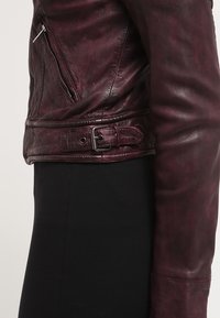 Giacca in pelle borgogna con un design aderente, caratterizzata da una tasca laterale con zip, dettaglio con fibbia in vita e superficie liscia.