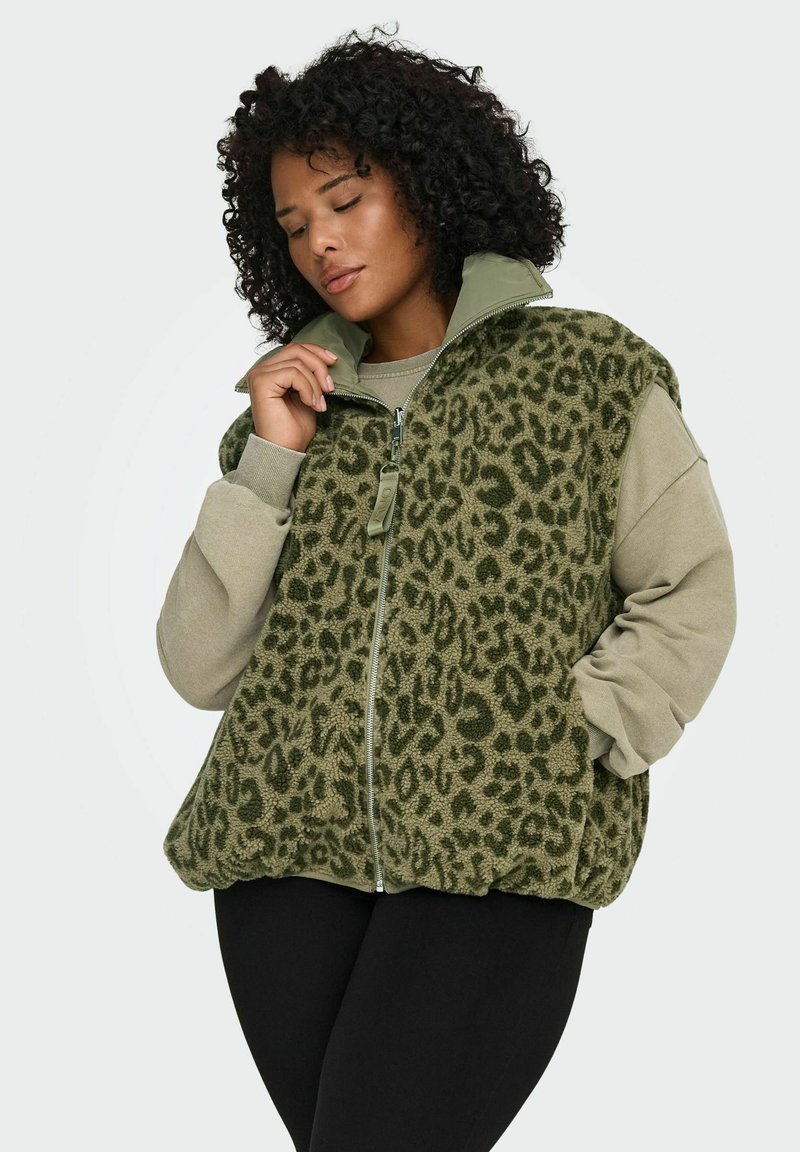 Woman with curly hair wearing a green leopard-print fleece vest over a beige sweatshirt, with one hand adjusting the vest's collar.