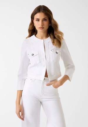 Frau mit welligem braunem Haar, die goldene Creolen, eine weiße cropped Jacke mit Vordertaschen und hoch taillierte weiße Hosen trägt, Hand in der Tasche.