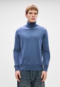 Pullover azul de gola alta em malha suave, com um colarinho e bainha canelados, apresentando um pequeno patch de logotipo no peito. Usado com jeans de denim.