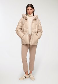 Giubbotto puffer beige con cappuccio, caratterizzato da un design trapuntato e tasche laterali. Indossato sopra un maglione leggero e abbinato a pantaloni chiari e sneakers.