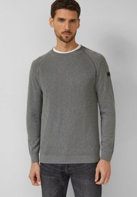 Uomo che indossa un maglione grigio aderente a coste sopra una maglietta bianca e jeans grigio scuro, in piedi contro uno sfondo chiaro e semplice.