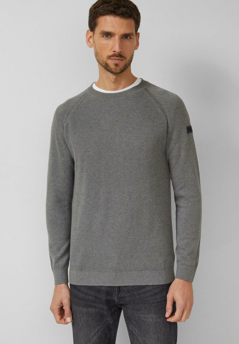 Uomo che indossa un maglione grigio aderente a coste sopra una maglietta bianca e jeans grigio scuro, in piedi contro uno sfondo chiaro e semplice.