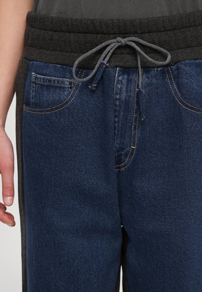 Joggers de mezclilla con una cinturilla elástica gris, cordón ajustable, bolsillos delanteros y costuras en contraste sobre la tela azul oscura.