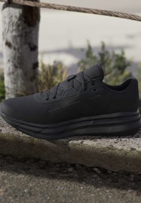 Baskets de sport noires avec dessus texturé, lacets et semelle épaisse et rembourrée reposant sur une surface en pierre à l'extérieur, près d'une corde et d'un tronc d'arbre.