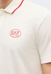 Λευκό κοντομάνικο πόλο με κόκκινη επένδυση στο γιακά και κόκκινο λογότυπο EA7 Emporio Armani στο στήθος.
