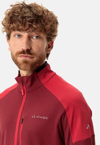 Giacca rossa con zip in tessuto testurizzato, caratterizzata da spalle in contrasto e un logo. Il colletto è alto con una chiusura a zip.