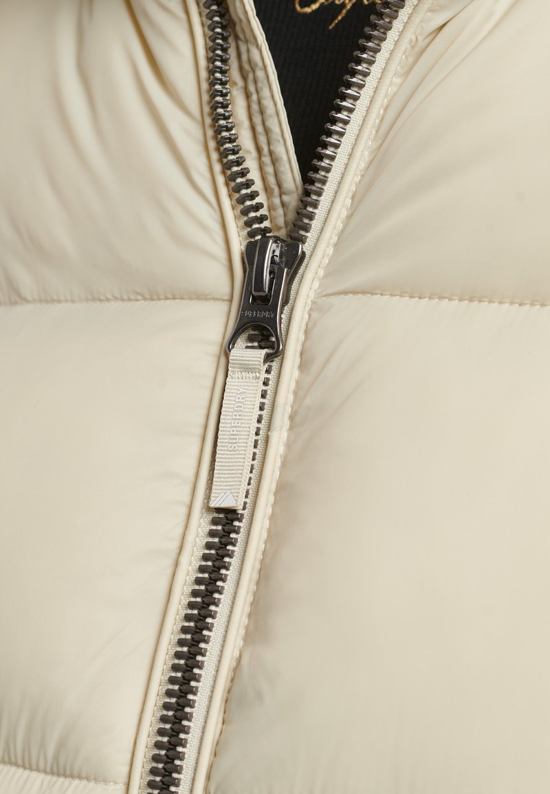 Giacca imbottita beige con zip argentata visibile. Il tirante della zip presenta un'etichetta chiara che riporta il marchio "Superdry."