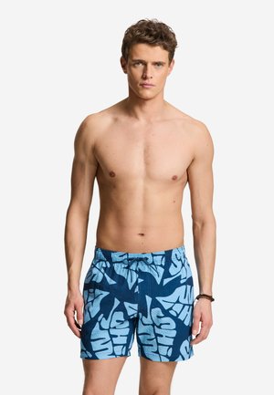 Jeune homme aux cheveux bruns courts portant un short de bain bleu avec un motif abstrait de lettres bleu clair, debout sur fond blanc.
