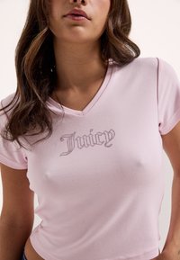 Γυναίκα που φοράει ένα ανοιχτό ροζ T-shirt με V λαιμόκοψη και τη λέξη "Juicy" με μικρές στρας στο στήθος.