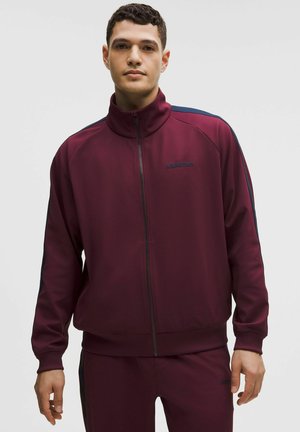 Ung mand iført en bordeaux fuld-zip jakke og matchende bukser med mørke sidestriber, stående foran en ensfarvet lys baggrund.