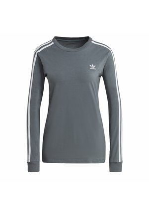 Personne portant un t-shirt Adidas gris, un blazer à carreaux gris posé sur les épaules, un jean gris, des baskets Adidas blanches et des chaussettes blanches avec des rayures noires.