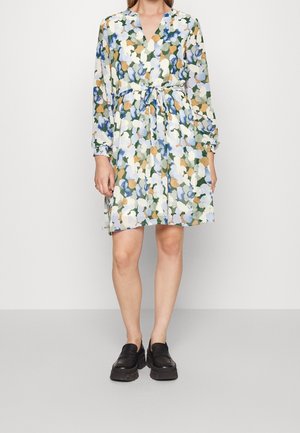 Kvinde iført knælængde, langærmet kjole med blåt, grønt, hvidt og beige blomsterprint og sorte kraftige loafers.