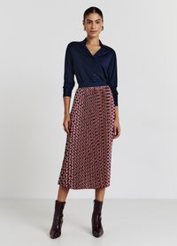 Chemise bleu marine boutonnée avec manches longues, associée à une jupe midi rouge à motifs circulaires. Le mannequin porte des bottines à talons foncées.