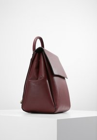 Sac à main en cuir rouge foncé avec poignée supérieure et fermeture à rabat, présenté debout sur un socle blanc contre un fond uni.