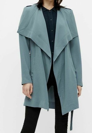 Femme portant un manteau bleu ardoise à ouverture devant avec larges revers sur une chemise noire boutonnée et un pantalon noir sur fond uni.
