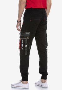 Schwarze Cargo-Hosen mit elastischen Bündchen, mehreren Taschen, silbernen Nieten und Reißverschlüssen. Mit roten Nähten und weißem Grafiktext.