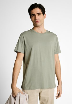 Jonge man met donker haar, gekleed in een lichtgroen T-shirt met korte mouwen en beige broek, houdt een beige jas in zijn linkerhand vast.
