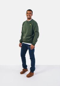 Grøn sweatshirt i bomuld med ribbede manchetter, et lille gult logo, parret med mørkeblå bukser og brune ruskinds sko. Hvid T-shirt synlig.
