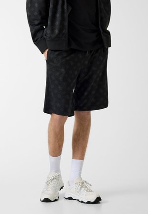 Mann trägt schwarze gemusterte Shorts, eine passende schwarze Jacke, weiße Sportsocken und weiße klobige Sneakers, steht mit den Händen in den Taschen.