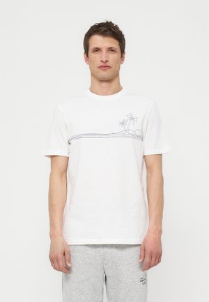 Άντρας που φοράει λευκό μπλουζάκι με λεπτή μαύρη γραμμή τέχνης φοίνικα και ήλιο, σε συνδυασμό με ανοιχτό γκρι αθλητικό παντελόνι, στέκεται μπροστά σε λευκό φόντο.