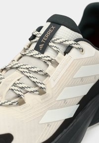Μπεζ αθλητικό παπούτσι με υφή στο πάνω μέρος, λευκές λεπτομέρειες, μαύρη καουτσούκ σόλα και χαρακτηριστικά κορδόνια με ασπρόμαυρο μοτίβο.