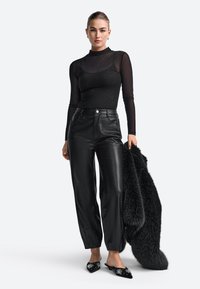 Top lung cu maneci lungi din material translucid negru cu strălucire subtilă, asociat cu pantaloni negri din piele ecologică cu talie înaltă. Modelul ține în mână o haină de blană neagră. Mules-uri negre cu broderie.